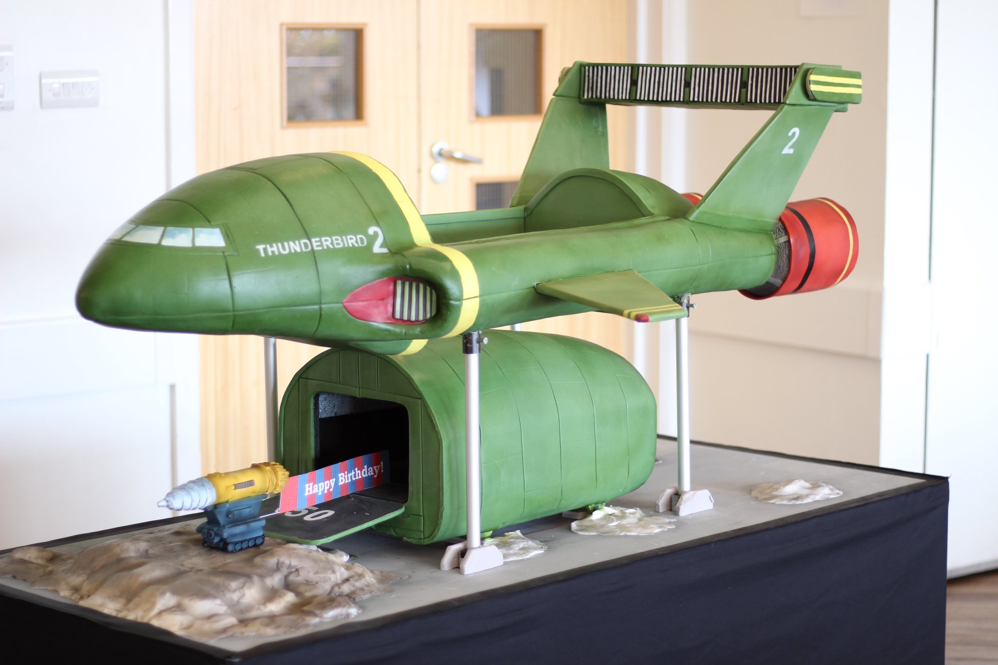 This is a side view of the Thunderbird 2 Cake. The frame of the cake has raised and a vehicles is coming out.